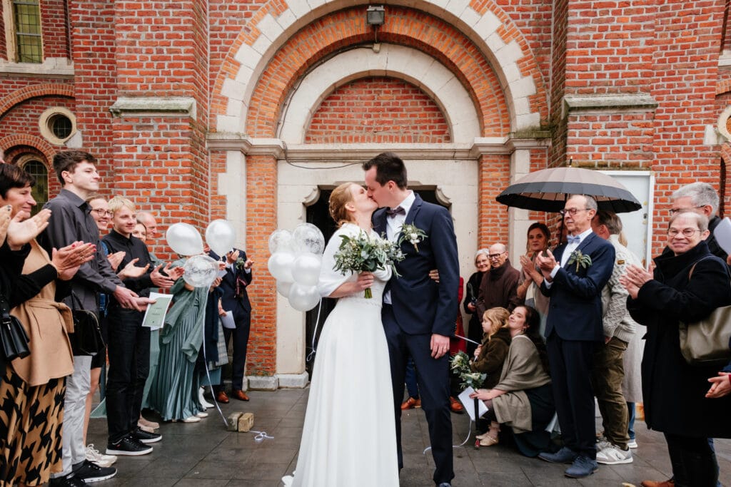 Huwelijksfoto van bruidspaar die elkaar kust bij buitenkomen van de kerk in Linden
